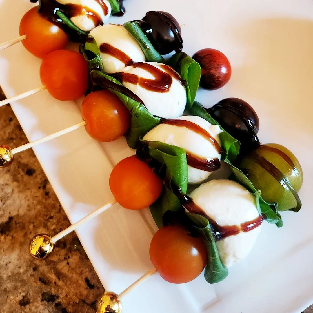 Caprese skewers with mozzarella, cherry tomatoes, basil, olives, and balsamic glaze on a serving platter.