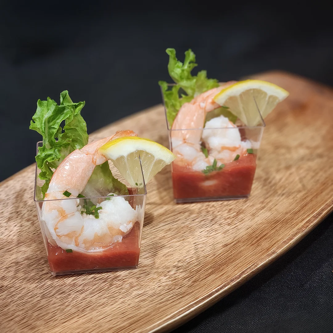 Individual shrimp cocktail cups with lemon wedges, cocktail sauce, and fresh greens on a serving board.
